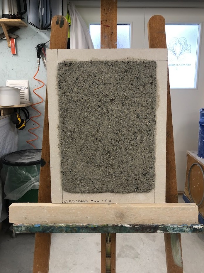 En bit gipslate med gipspuss på. Gipsplaten er plassert på et staffeli.